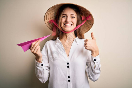 戴着亚洲传统帽子的年轻漂亮红发女人拿着纸飞机开心地笑着做好手势图片