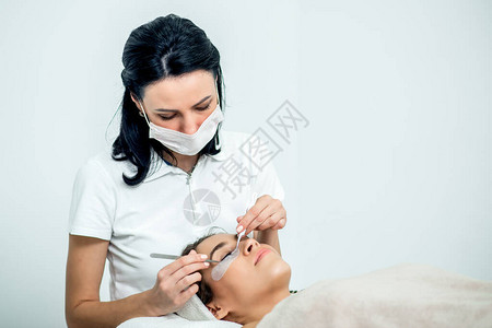 Beautician工作在延长眼睫毛程序期间病人的白色背景图片