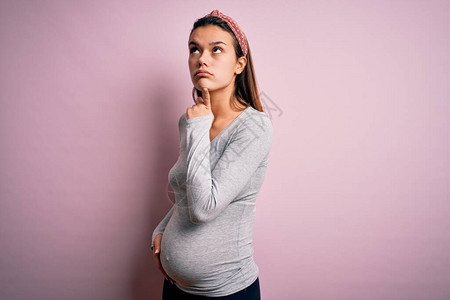 怀着与世隔绝的粉红背景的怀孕年轻少女预产婴儿想着用手指在下巴上的疑惑图片