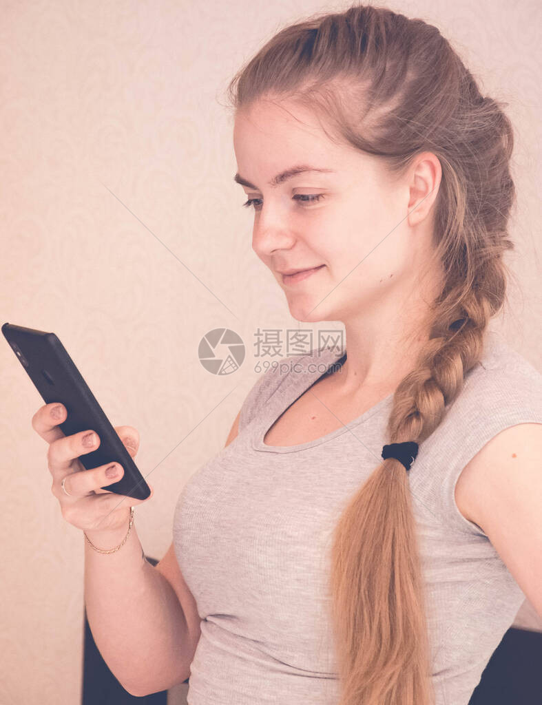 穿着灰色T恤和长的大辫子的年轻漂亮女孩正在研究智能手机拿着镰刀的女孩在社交图片