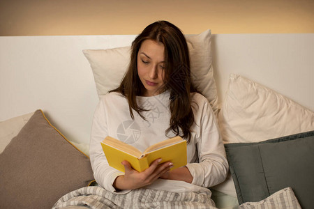 穿着睡衣的漂亮黑发女孩在懒惰的一天坐在床上看书图片