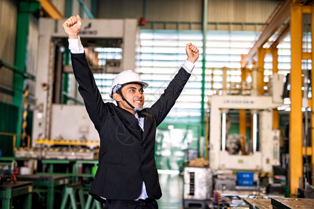 戴着防护白色安全帽的快乐微笑商人举手庆祝他的工厂铁备件的新高销量亚洲人在重工业中取得成功的画图片