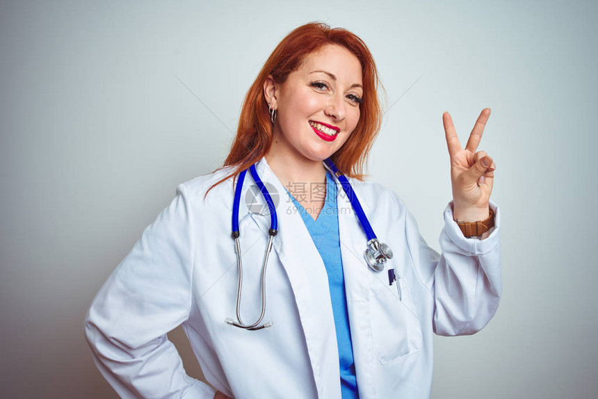 年轻红发女医生在白色孤立的背景上使用听诊器微笑地看着摄影机手举起来做胜利标志第图片