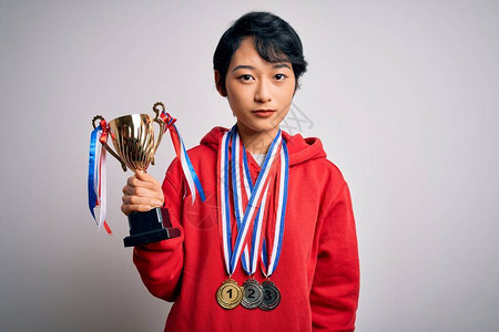 年轻美丽的亚洲女孩赢家拿着奖杯身着白背景的奖牌自信地表现在聪明的面孔图片