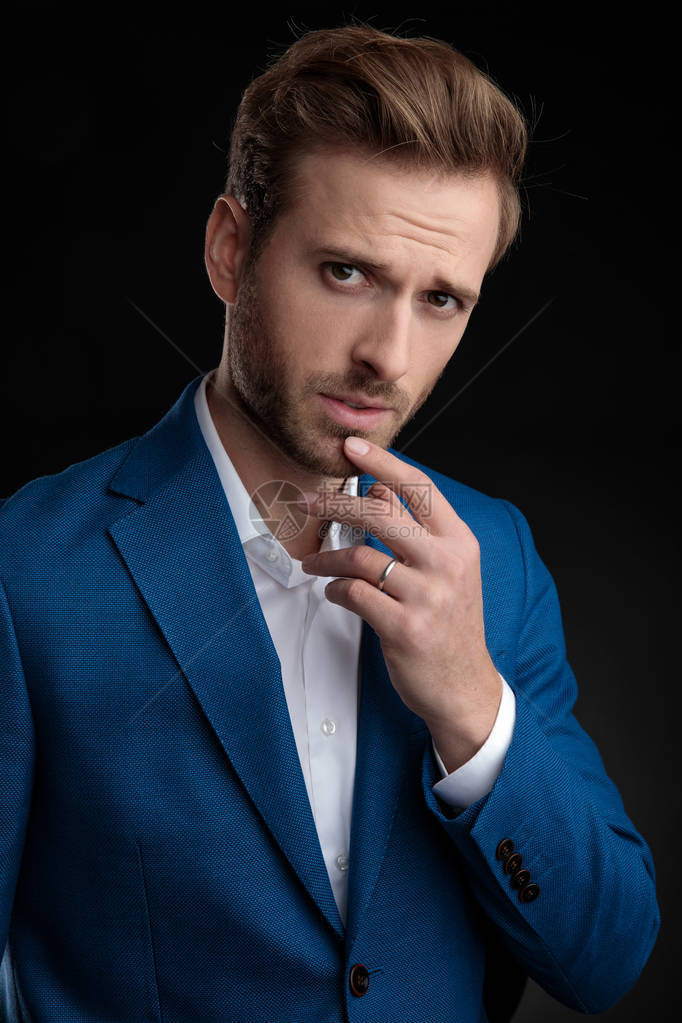 身穿蓝西装站在黑工作室背景上站着的可疑男子举起手顶下巴图片
