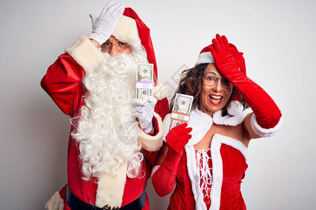 穿着圣诞老人服装的老夫妇拿着美元在孤立的白色背景上惊讶地用手捂着头图片