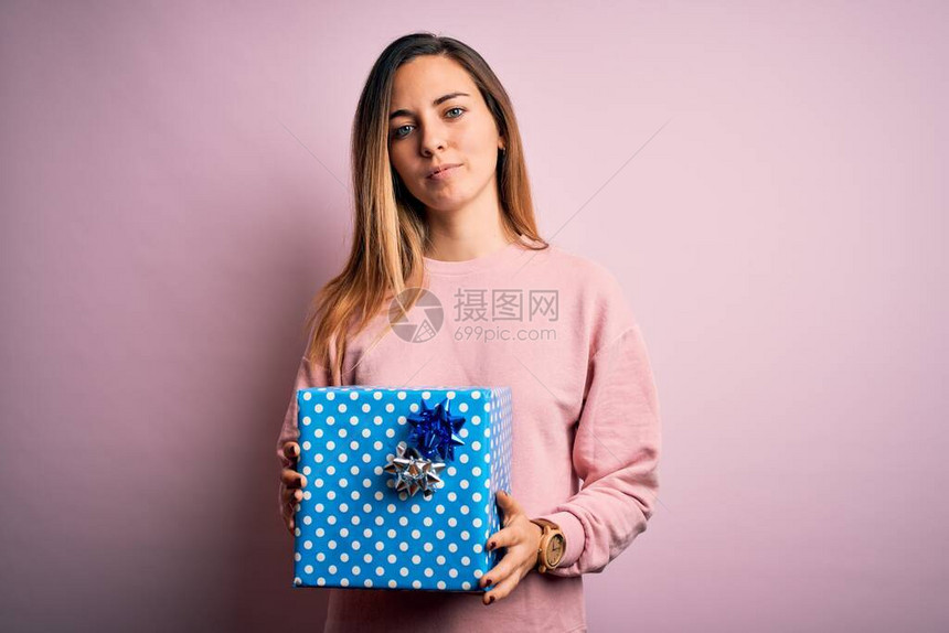 蓝眼睛的年轻金发美女在粉红背景上拿着生日礼物自信地表现在聪明的面孔图片