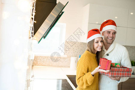 穿着红色帽子的年轻男女在厨房里拿着圣诞礼物站着时图片