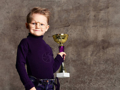 穿着牛仔裤毛衣和眼镜的聪明小男孩在混凝土墙的背景下庆祝他赢得的金奖杯图片