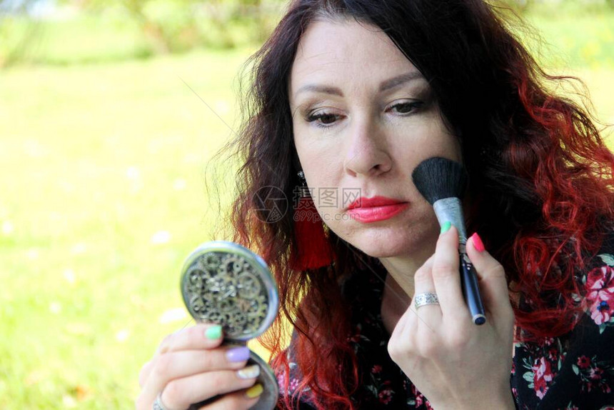一个红色卷发的女人在绿色的草坪上纠正她的妆容用刷子把粉末放在小镜子里用化妆刷涂图片