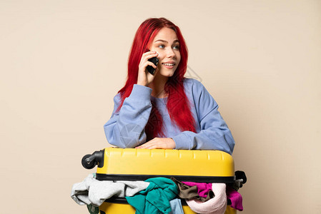 旅行女孩带着一个手提箱满装衣物的衣服孤立在蜜蜂背景上与某人和手机交谈图片