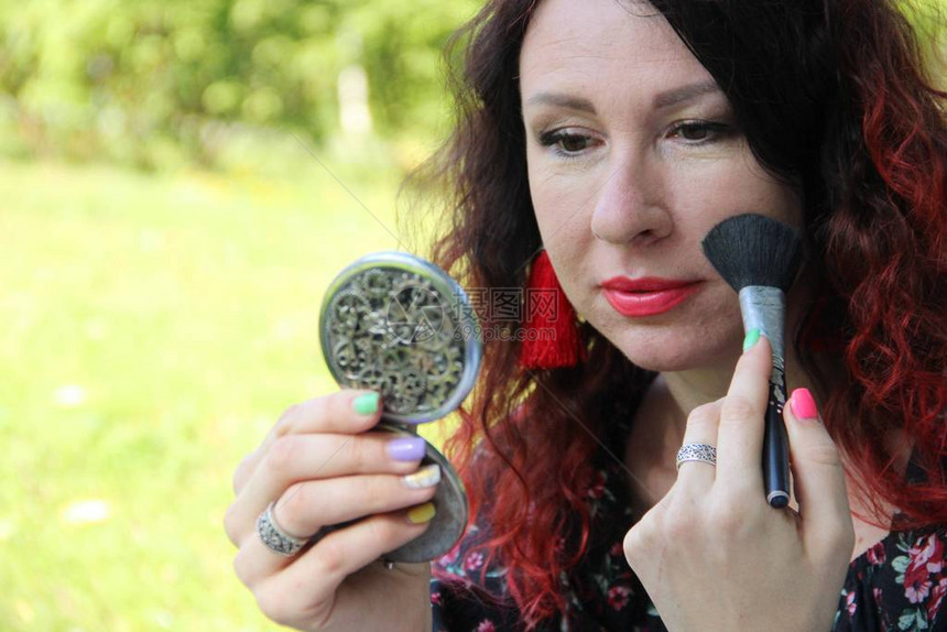 用化妆刷涂抹粉底或腮红一个红色卷发的女人在绿色的草坪上纠正她的妆容用刷子把粉末图片