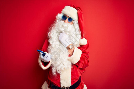 穿着圣诞老人服装的老人在孤立的红色背景上用智能手机严肃地思考问题图片