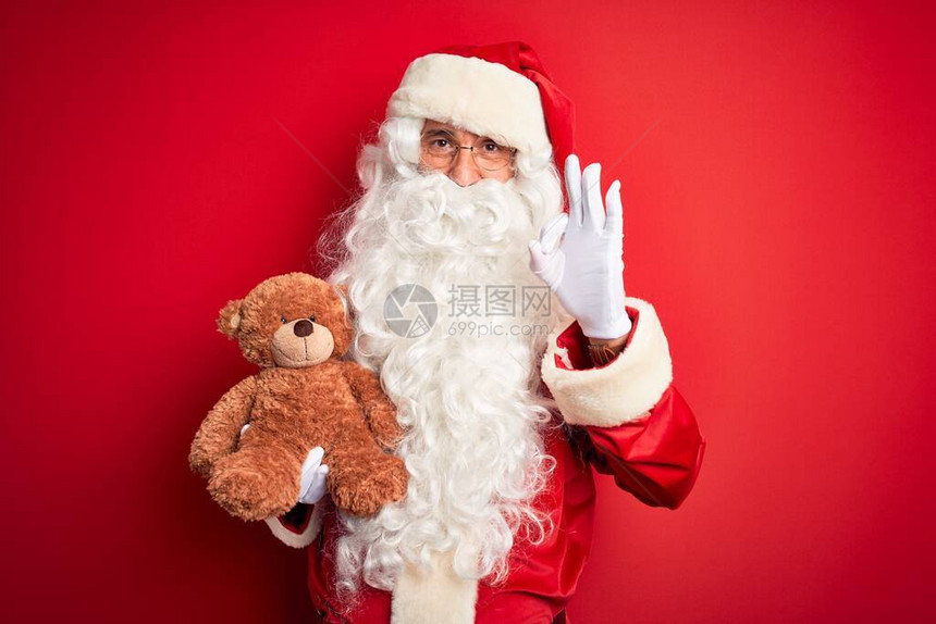 身着圣诞老人服装的中年男子带着泰迪熊在孤立的红色背景上举着特迪熊用图片