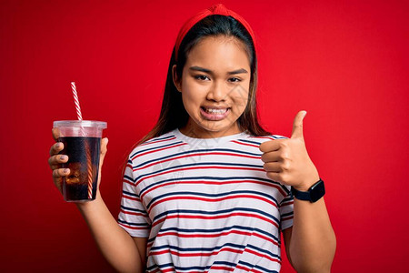 年轻的亚洲女孩在孤立的红色背景上用稻草喝可乐汽水图片