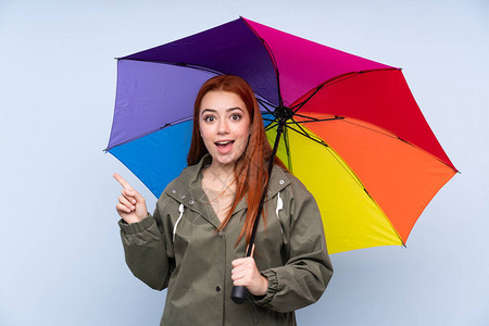 带着伞的红头少女在孤立的蓝色背景上拿着雨伞图片