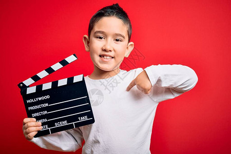 小男孩在孤立的红色背景上拿着电影导演隔板拍摄视频图片