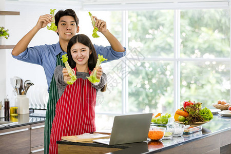 亚洲夫妇在围裙里做沙拉卷展示给大家看蔬菜和笔记本放在柜台上在厨房图片