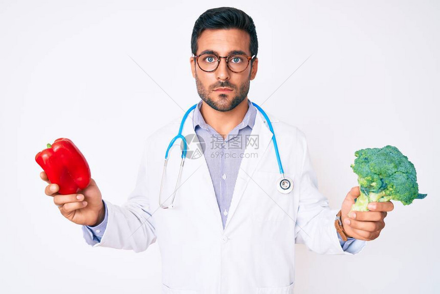 身着医生听诊器手持红胡椒和花椰菜的年轻西班牙男子在脸上露出严肃的表情图片