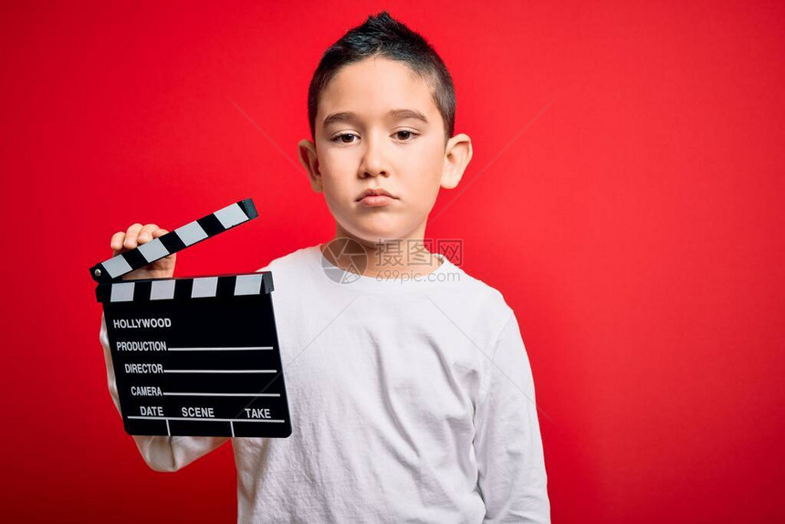 小男孩在孤立的红色背景上拿着电影导演隔板拍摄视频图片