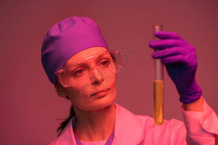 穿着白大衣防护手套和眼镜的年轻女实验室工作人员看着烧瓶中的图片