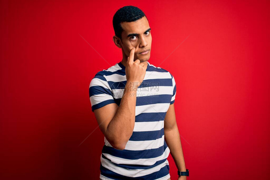 身穿轻便条纹T恤的非洲英俊男子站在红色背景上指着看你的手势可疑的表情图片