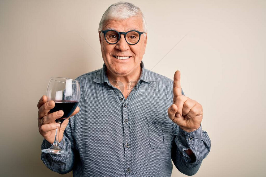 高级英俊修士在孤立的白种背景上喝着一杯红葡萄酒惊讶于一个想法或问题与快乐图片