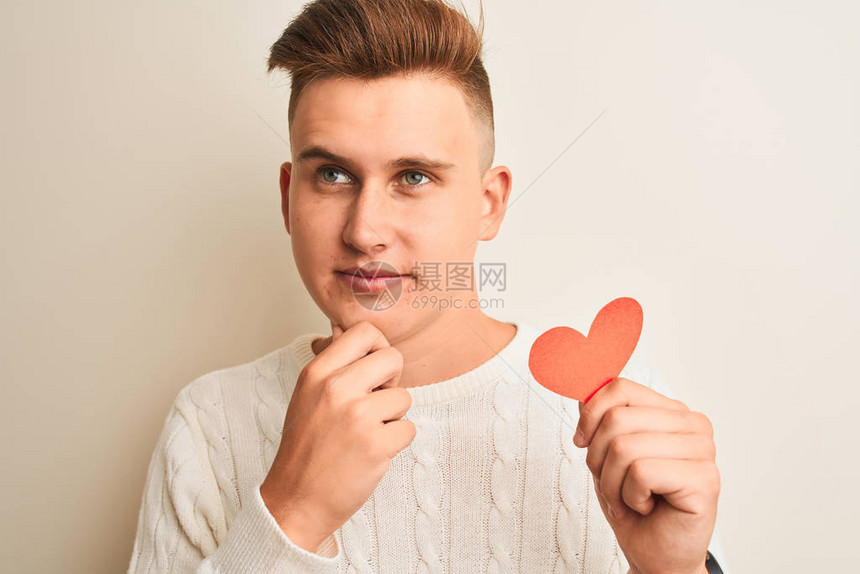 年轻英俊的浪漫男子拿着纸心站在孤立的白色背景上面对严肃的面孔思考问题图片