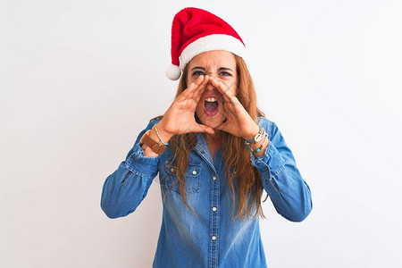 年轻美丽的红发美女戴着圣诞帽子在孤立的上喊着愤怒大叫用手图片