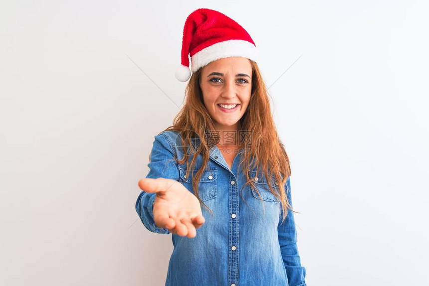 身戴圣诞帽的年轻红发美女在孤立的背景中微笑地欢笑着献出手掌来给予帮助和接受图片