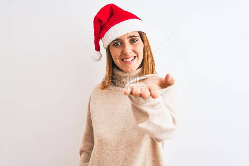 穿着圣诞帽的美丽的红头发女人在孤立的背景中微笑地欢笑着献出手掌来给予帮助和接图片