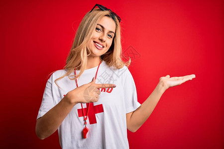 年轻美丽的金发救生员女人穿着t恤与红十字使用口哨惊讶并微笑着对镜头图片
