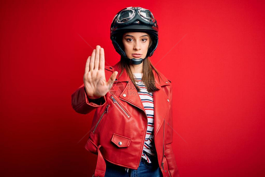年轻漂亮的黑发摩托车手女人戴着摩托车头盔和红色夹克做停止用手掌脸上带着消极和严肃的图片