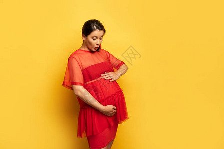 穿着红色束腰外衣的怀孕年轻女子在黄色上摸着肚子背景图片