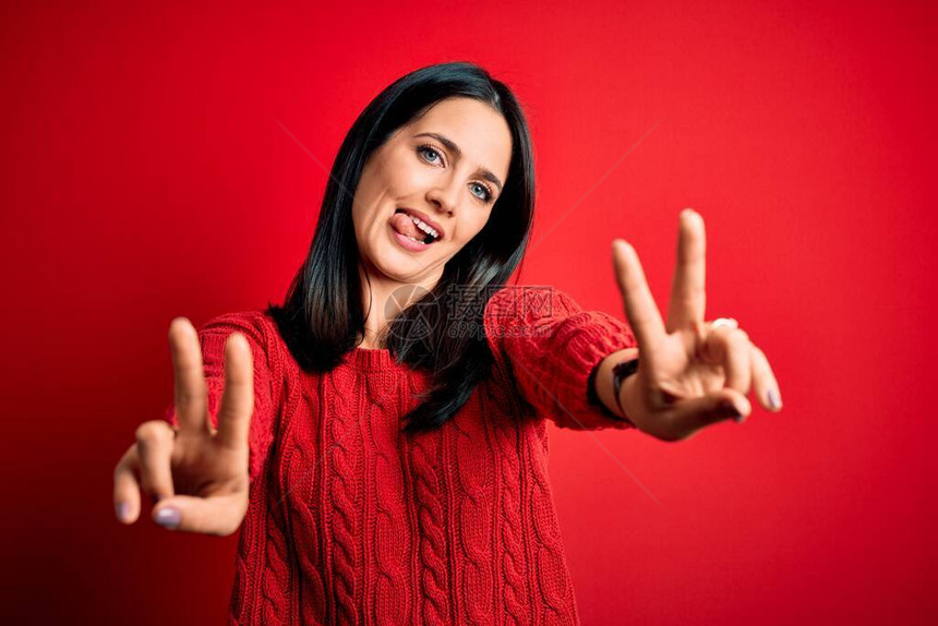 蓝眼睛的黑发年轻女人穿着随身毛衣在孤立的红色背景上微笑用舌头笑出两只手的指来做胜图片