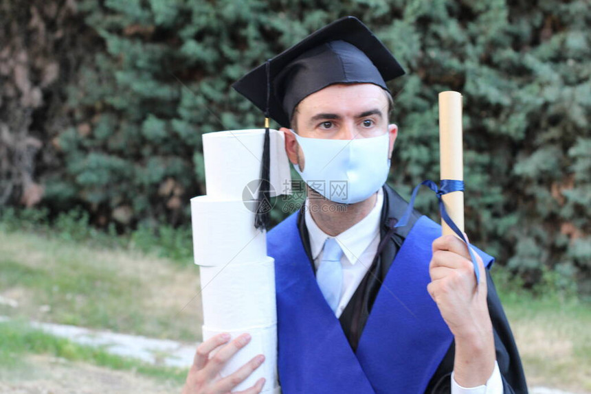 穿着毕业服装的英俊青年学生在公园持有滚版文凭和卫生纸卷的图片