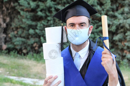 穿着毕业服装的英俊青年学生在公园持有滚版文凭和卫生纸卷的图片