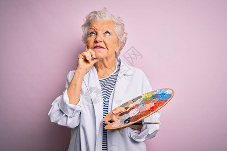 资深美丽的灰发艺术家女用画笔和调色板在粉红色背景上严肃地思考问题图片