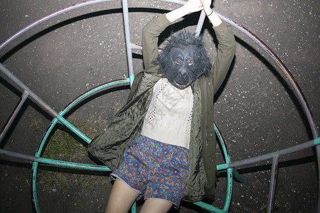 戴着大猩面具外套和短裤的滑稽白人女孩躺在儿童游乐场的金属结构上图片