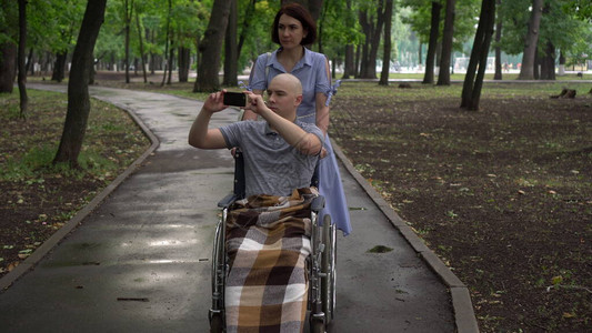 一名妇女带着一名坐在轮椅上的肿瘤科年轻男子穿过公园图片