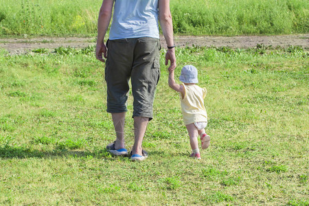 岁的小女孩牵着爸的手去父亲带着孩子穿过绿草婴儿在父母的支持下学会走路图片