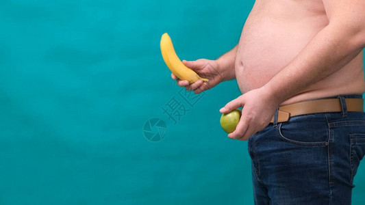 大肚子的胖子手里拿着一个青苹果和一根香蕉健康饮食和减肥图片