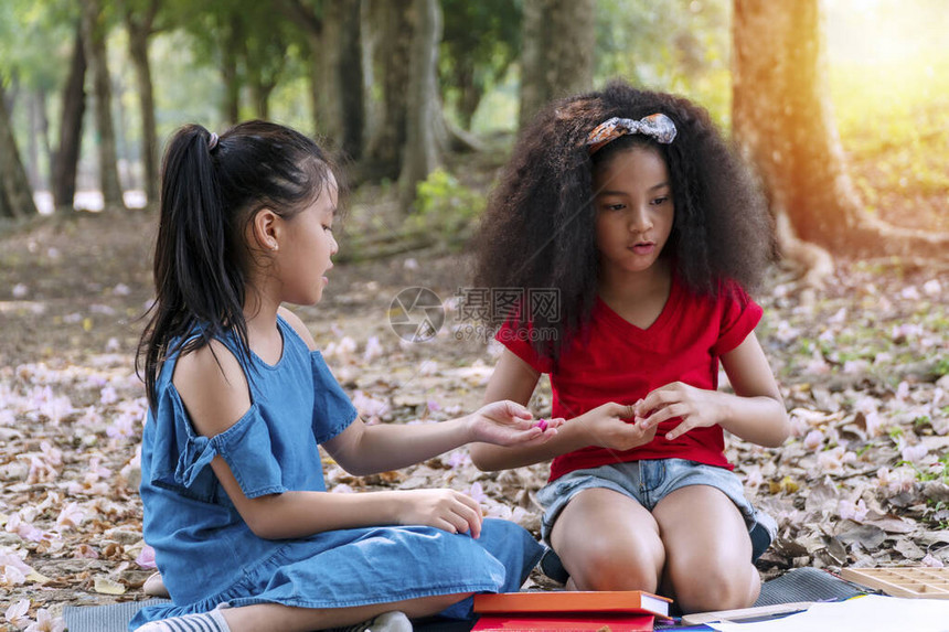 快乐的两个小女孩拿着橡皮泥在公园里坐在一起看东西与亚洲朋友一起玩时手里拿着橡皮泥的混血小学生图片