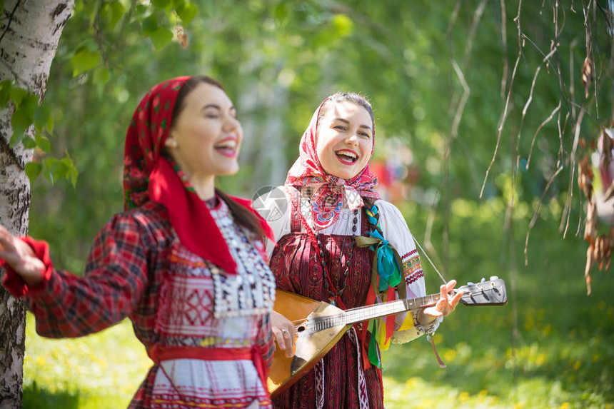 两个穿着传统俄罗斯服装的年轻有魅力女人在森林里唱歌其中一人在玩芭拉莱卡图片