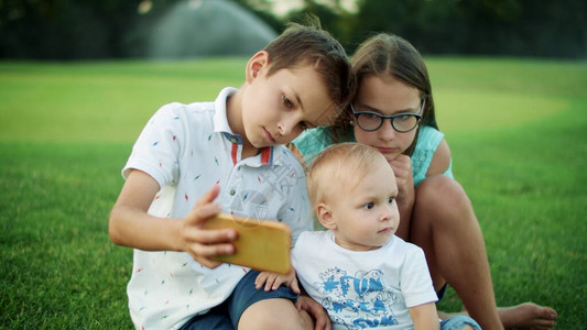 专注的孩子们坐在夏日草地上的绿草上可爱的男孩在绿色公园与姐和弟在手机上自拍照片可爱的孩子们在户外看图片