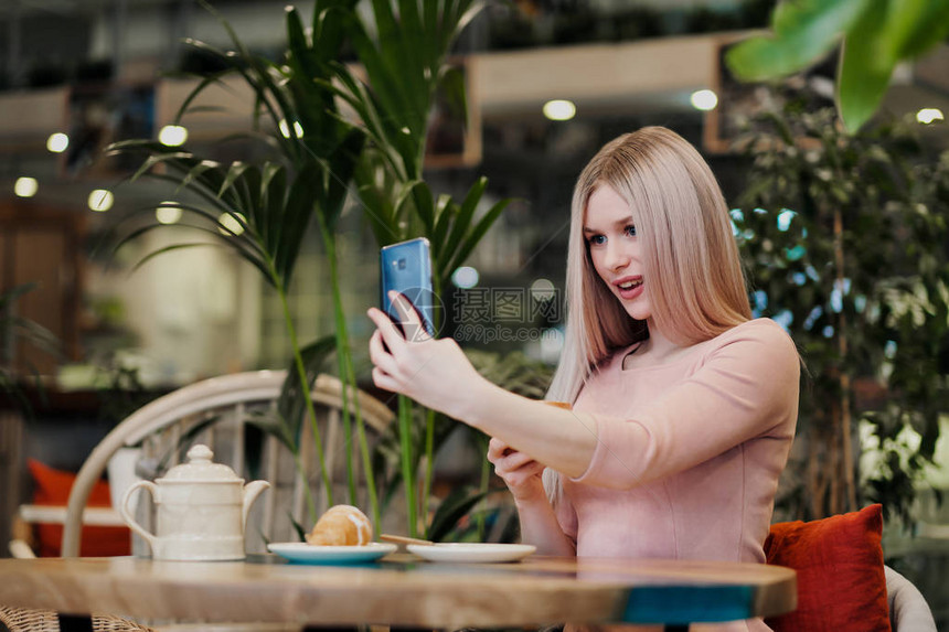 年轻漂亮的女孩坐在一张绿色的咖啡馆里用羊角面包喝茶聊天大笑和在智能手机上拍照与朋友交流图片