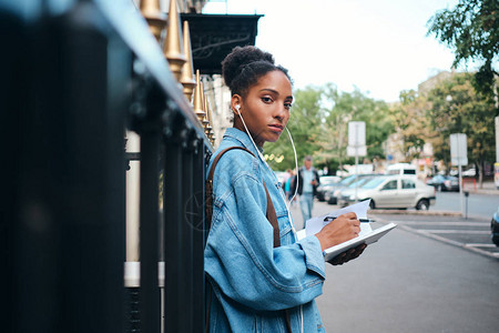 身穿牛仔衣的非裔美籍女学生带着笔记纸满怀自信地在市街背景图片