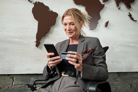 坐在椅子上并使用智能手机和信用卡在网上转账时进行汇款的成熟商业女企业背景图片