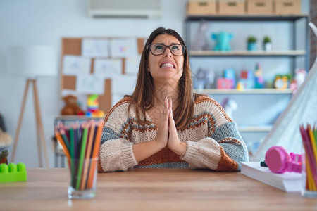 年轻漂亮的女教师穿着毛衣和眼镜坐在幼儿园的桌子上乞讨和祈祷图片