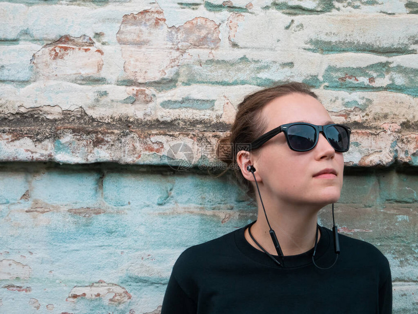 穿着黑色t恤和太阳镜在城市背景下戴着耳机听音乐的摇滚风格的年轻迷人女孩的画像在旧的蓝砖墙背景上的无图片